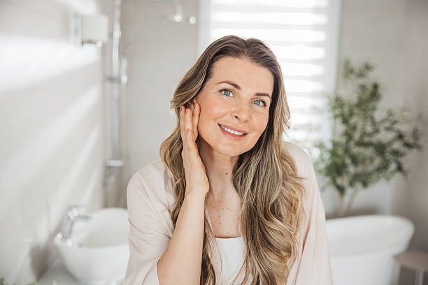 Mature woman applying moisturizer on face in front of mirror in bathroom